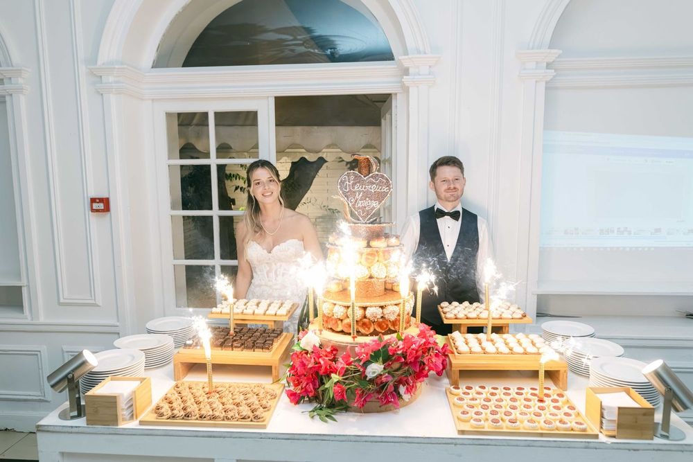 Bride and groom smiling behind elegant wedding dessert table with sparklers and floral arrangements.