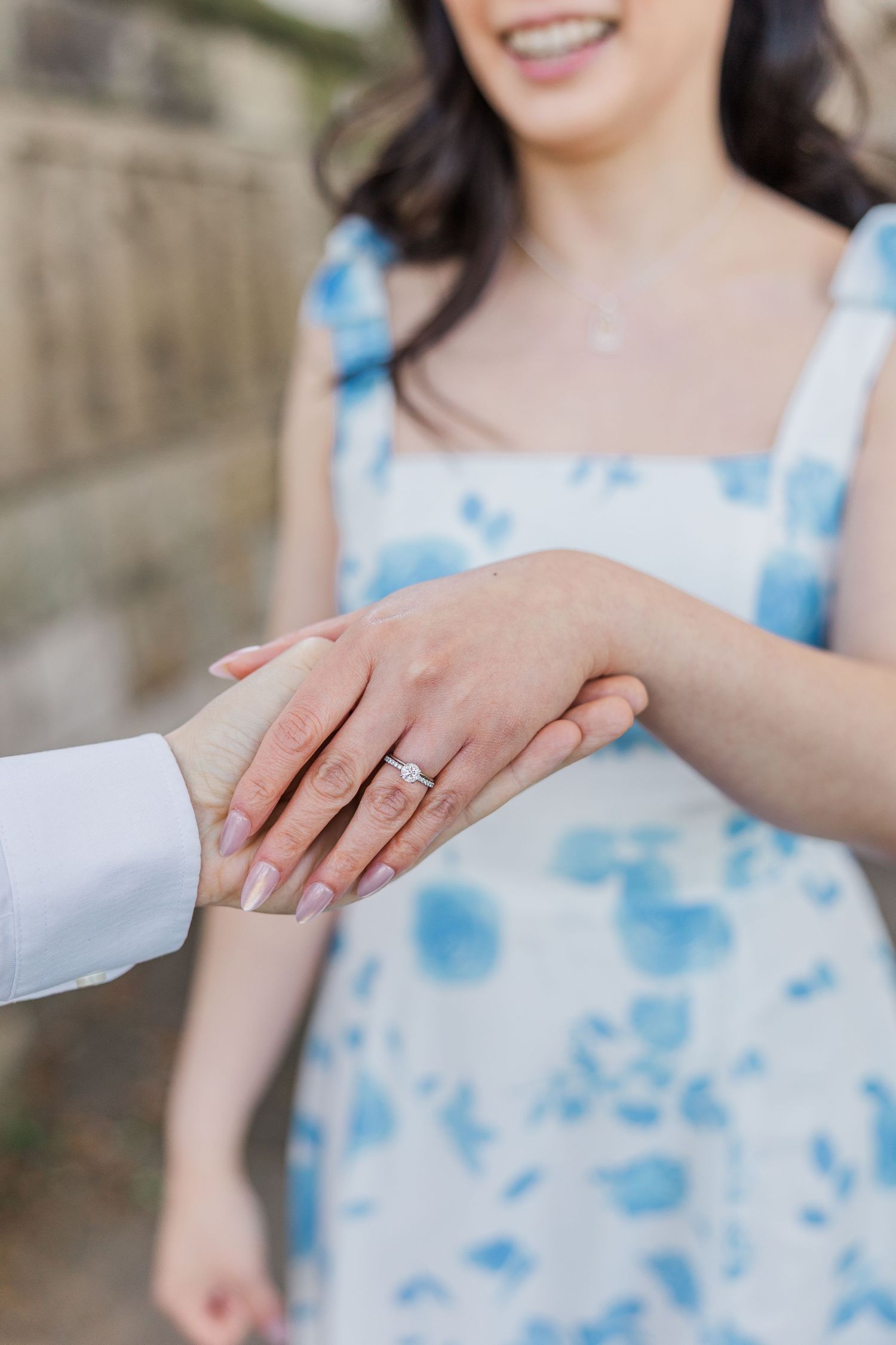Close-up of a woman in a blue floral dress smiling as her partner holds her hand displaying an engagement ring.