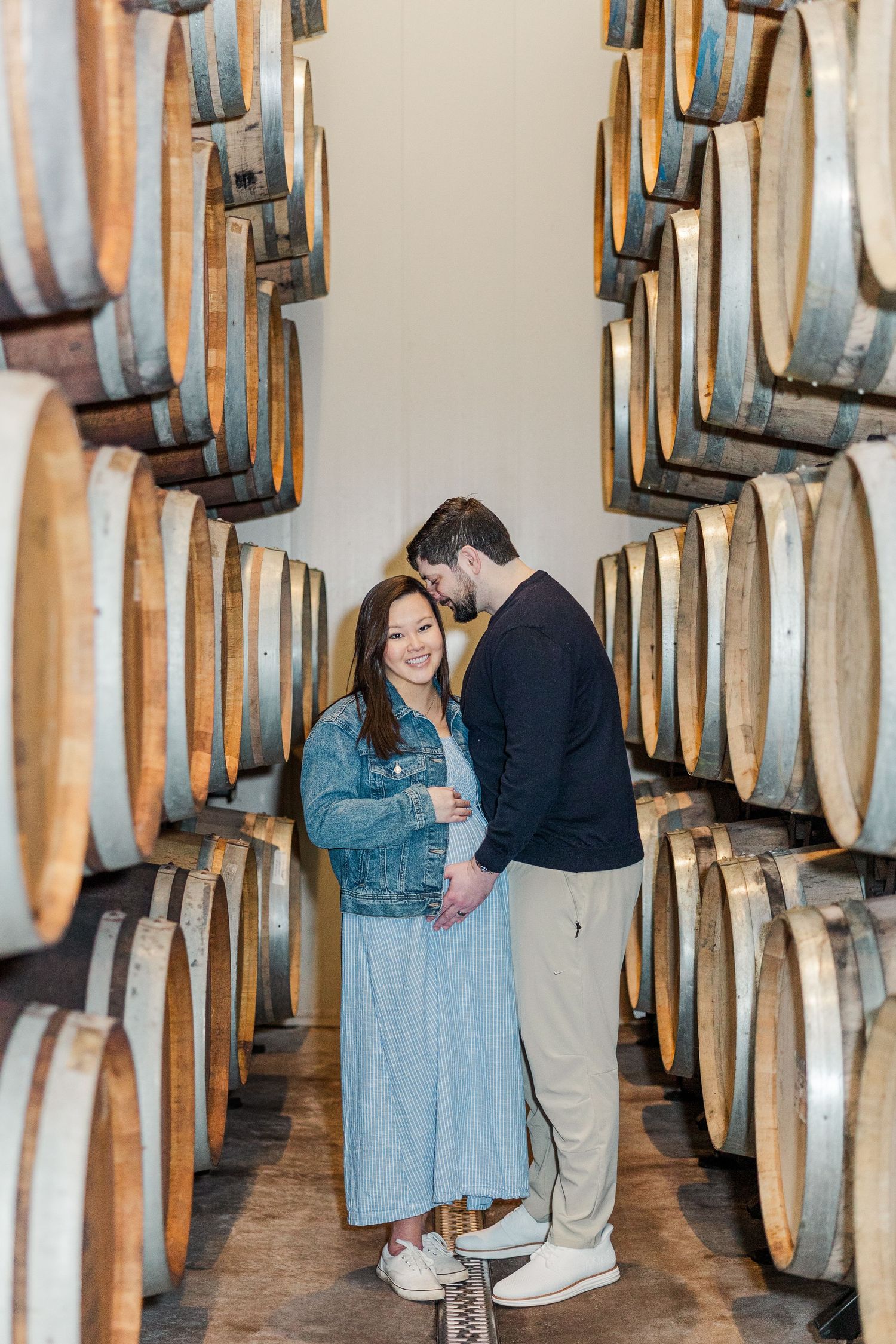 Couple poses between rows of wooden barrels in a winery cellar, woman in denim jacket and blue dress.