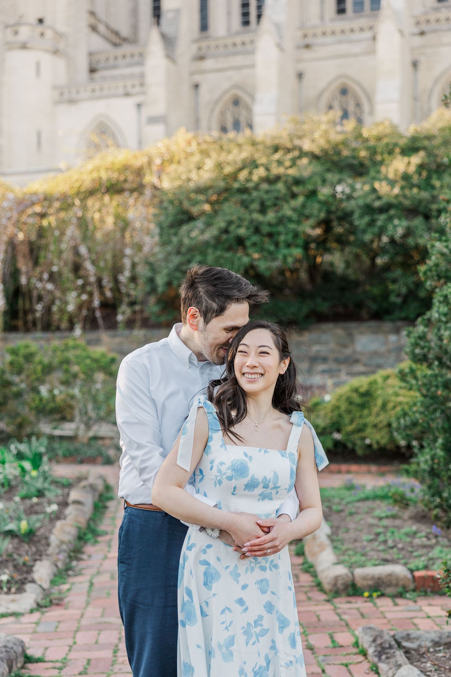 Couple embracing in garden near gothic cathedral, woman in floral dress smiling joyfully during engagement photo session.
