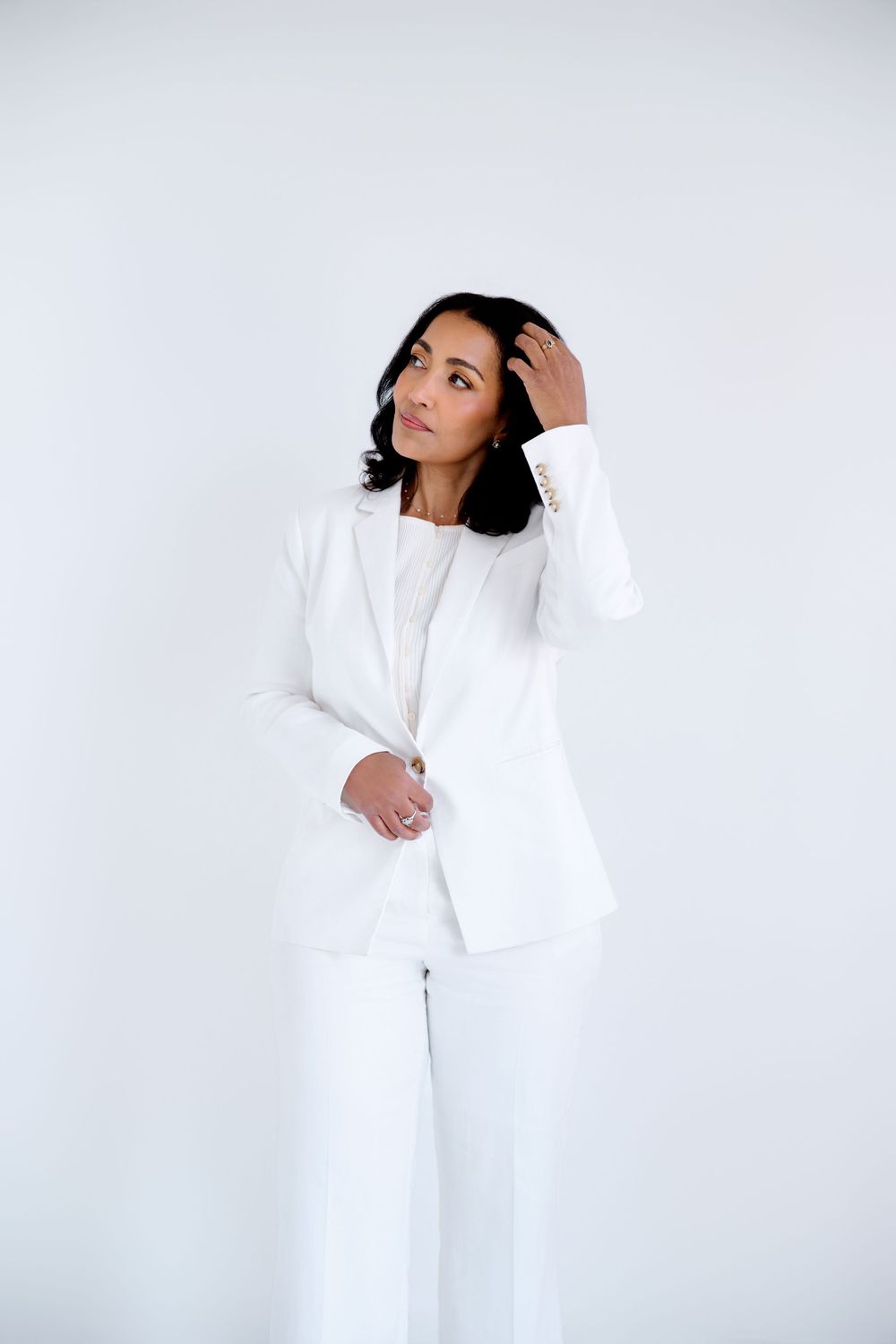 A confident woman in a white blazer and pants poses against a white background, touching her hair elegantly.