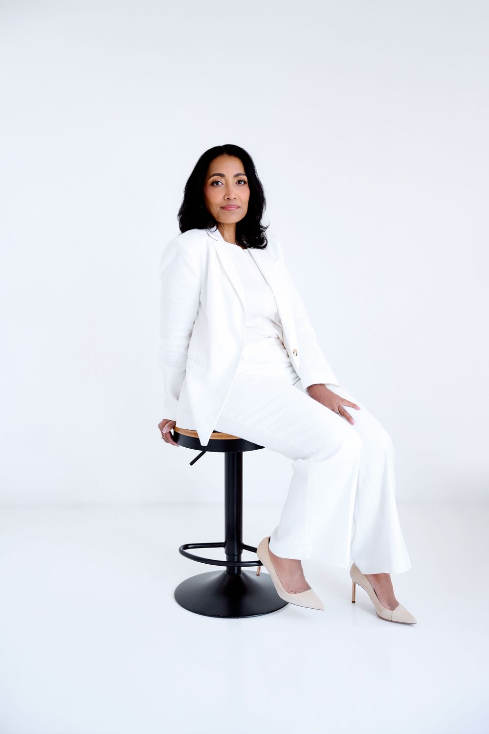 Woman in white suit seated on black bar stool against white background, posing confidently in heels.