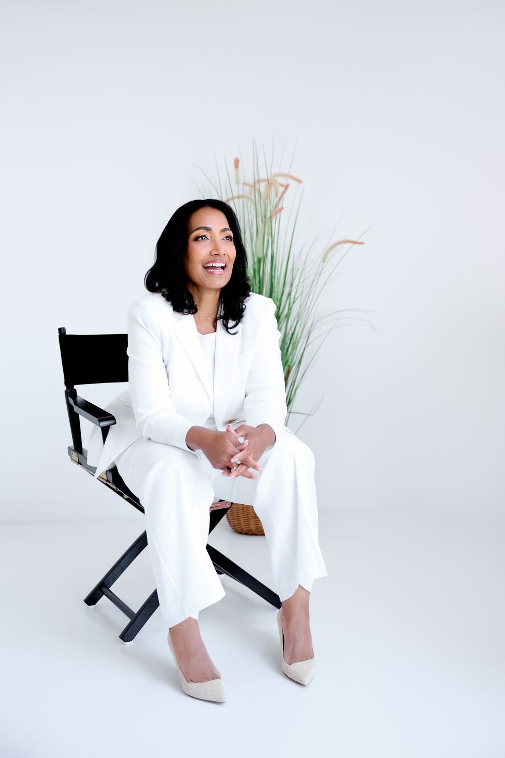 Woman in white suit sitting in director's chair, smiling confidently against a light background with decorative grass.
