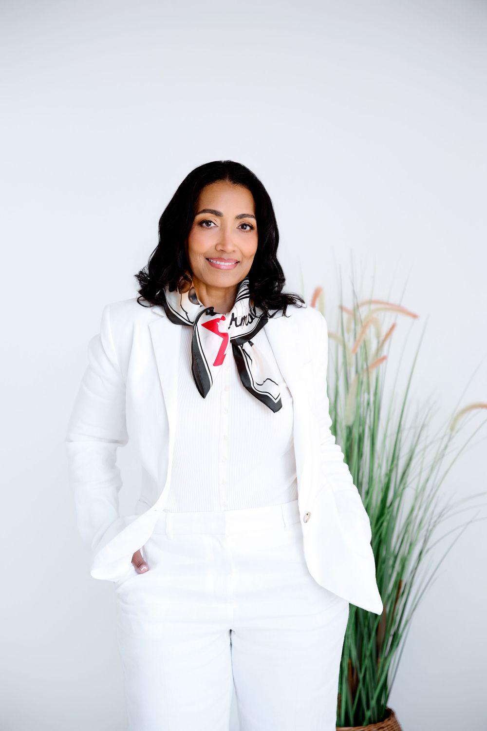 Woman in white blazer and pants with silk scarf smiling confidently in a professional studio setting.