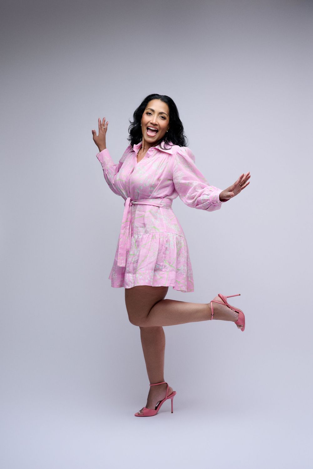 Woman in pink floral mini dress and heels posing joyfully with raised hands and one leg lifted on gray background.