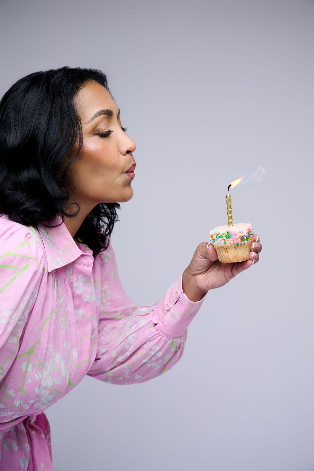 Woman in pink floral dress blowing out a birthday candle on a decorated cupcake against gray background.