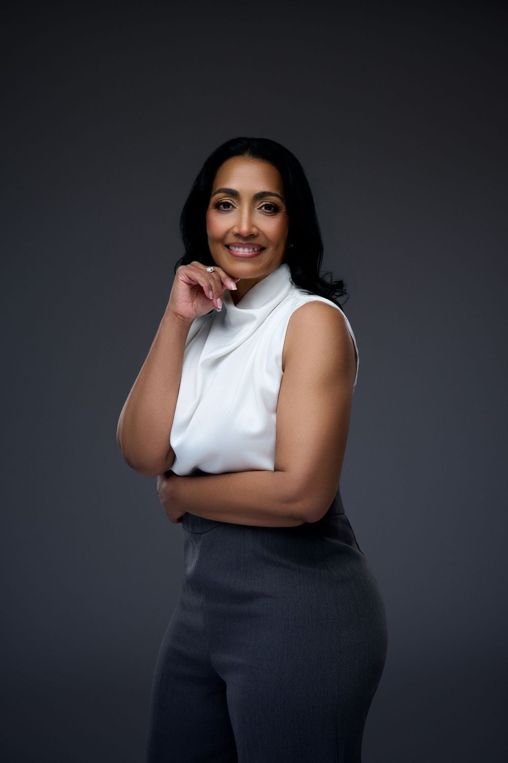 Professional woman in white sleeveless turtleneck and gray pants smiling confidently against dark background.