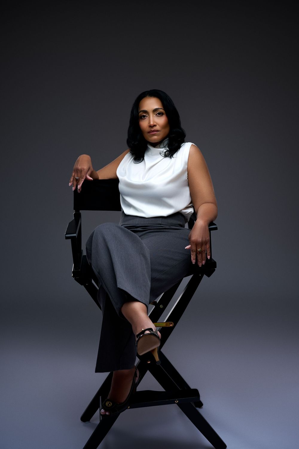 Woman in white sleeveless top and gray pants sits confidently in a director's chair against a gray background.