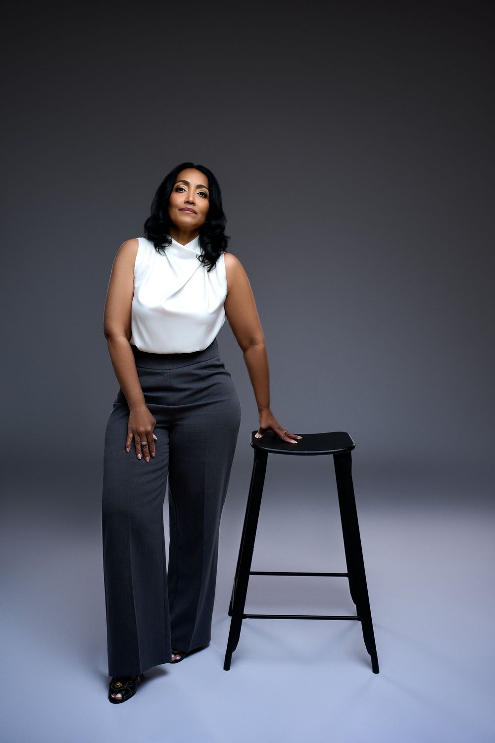 Woman in white sleeveless top and gray wide-leg pants poses beside a black bar stool in a studio.
