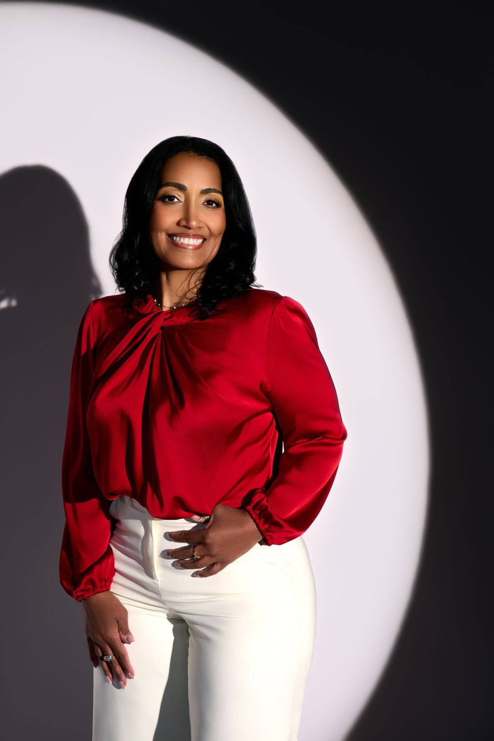 Smiling woman in red satin blouse and white pants poses confidently against dark background with spotlight.