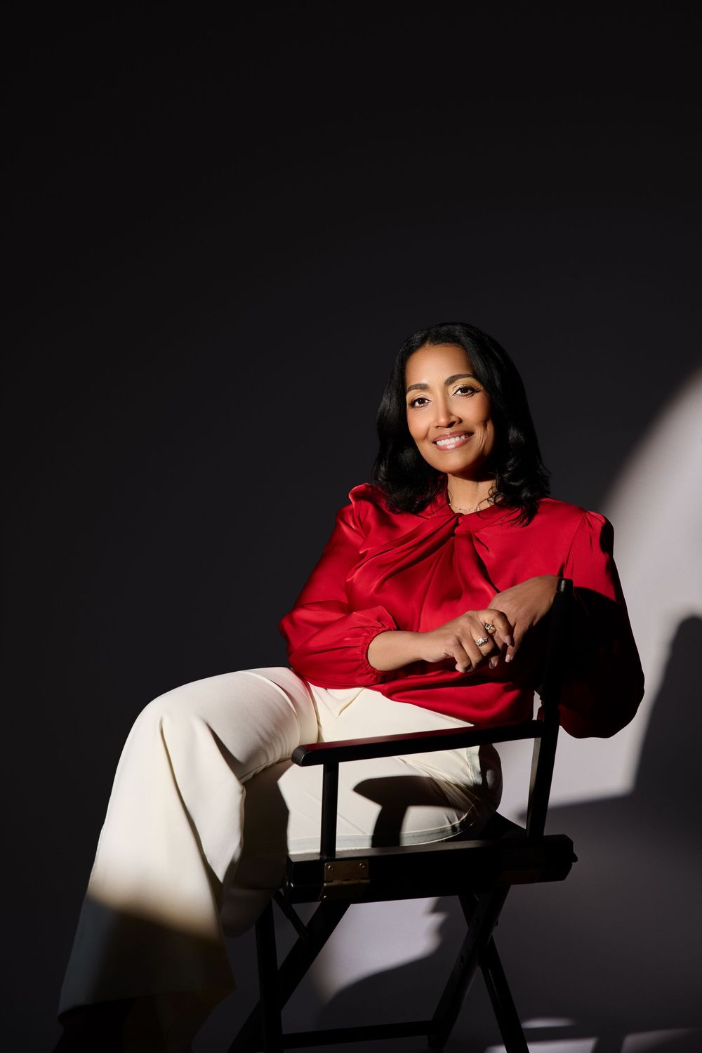 Woman in red blouse and white pants sitting in a director's chair, smiling against a dark background.