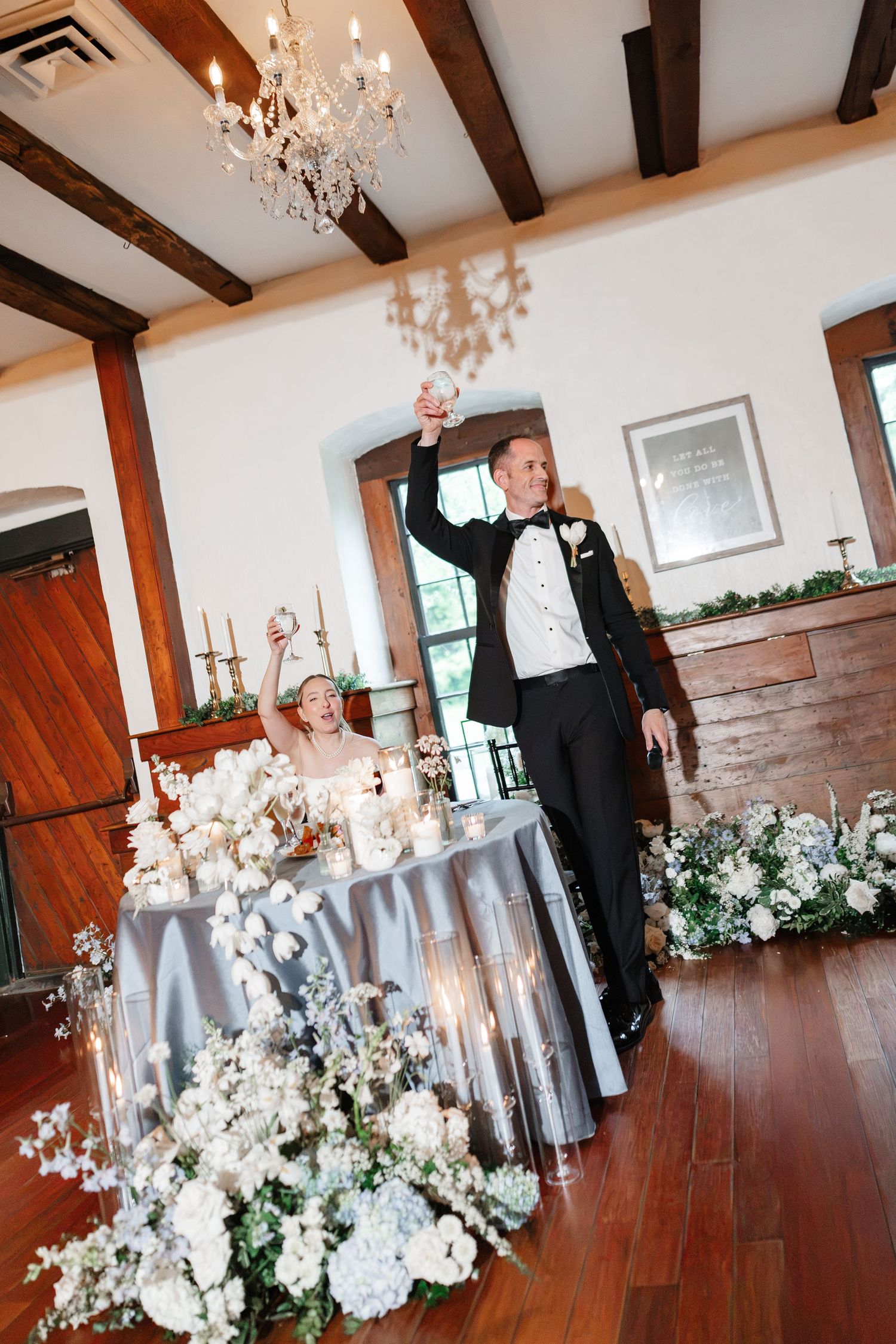 Bride and groom raising hands joyfully at elegant wedding reception with floral arrangements and chandelier.