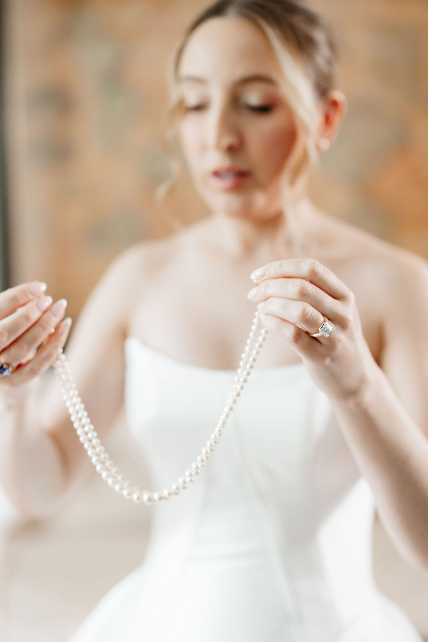 Bride in strapless white gown holds a delicate pearl necklace while wearing her diamond engagement ring.