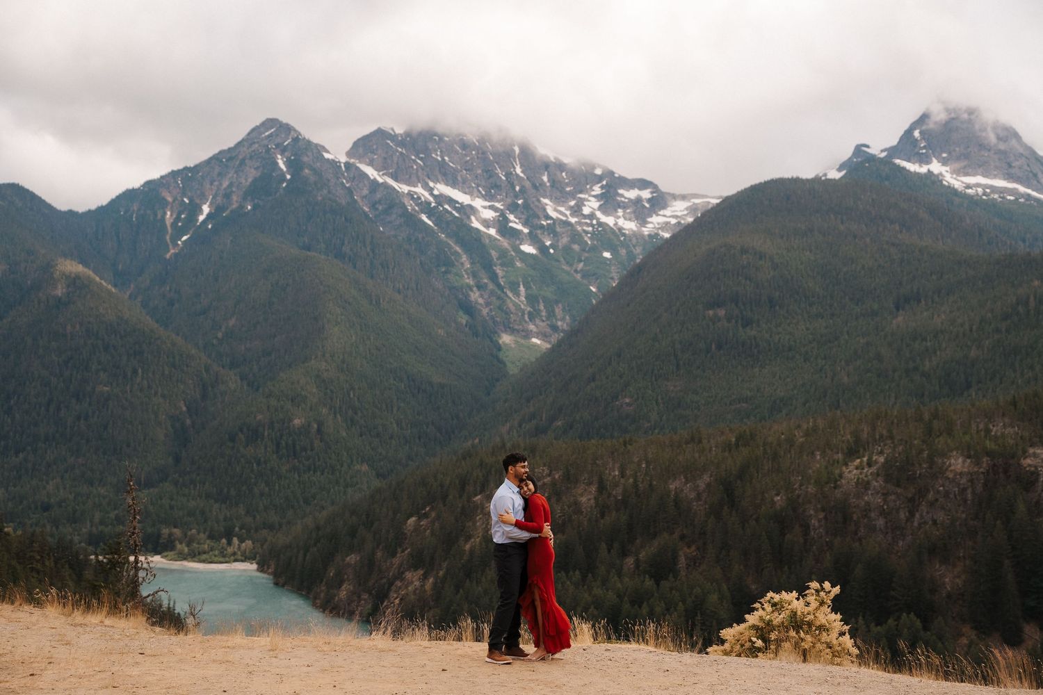 Couple embracing at scenic mountain overlook with river valley, evergreen forests, and snow-capped peaks at lake Diablo engagment.
