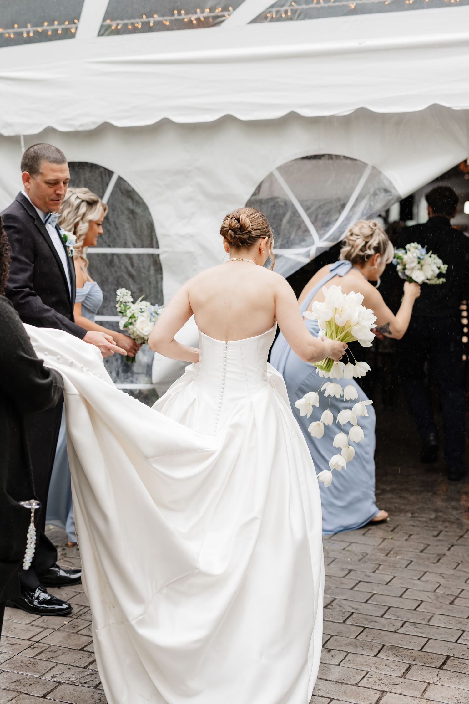 Bride in strapless white gown with train walking with bridesmaids in blue dresses carrying white tulip bouquets.