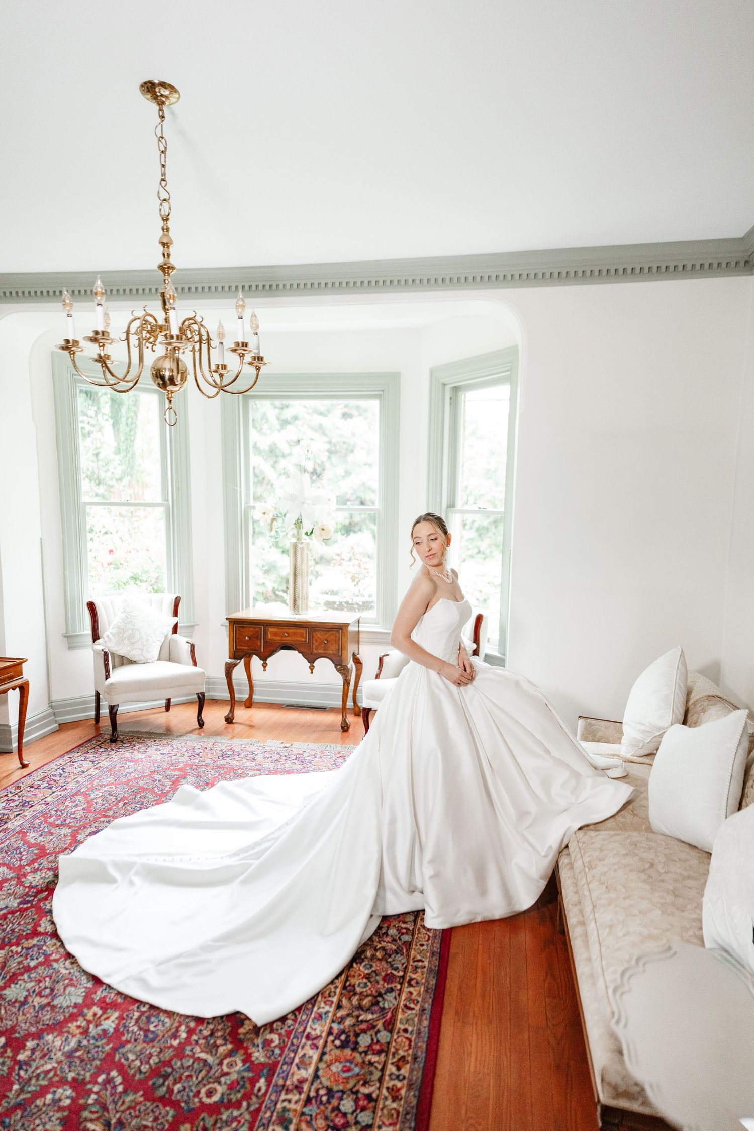 Bride in elegant white ball gown with long train poses in a classic room with chandelier and bay windows.