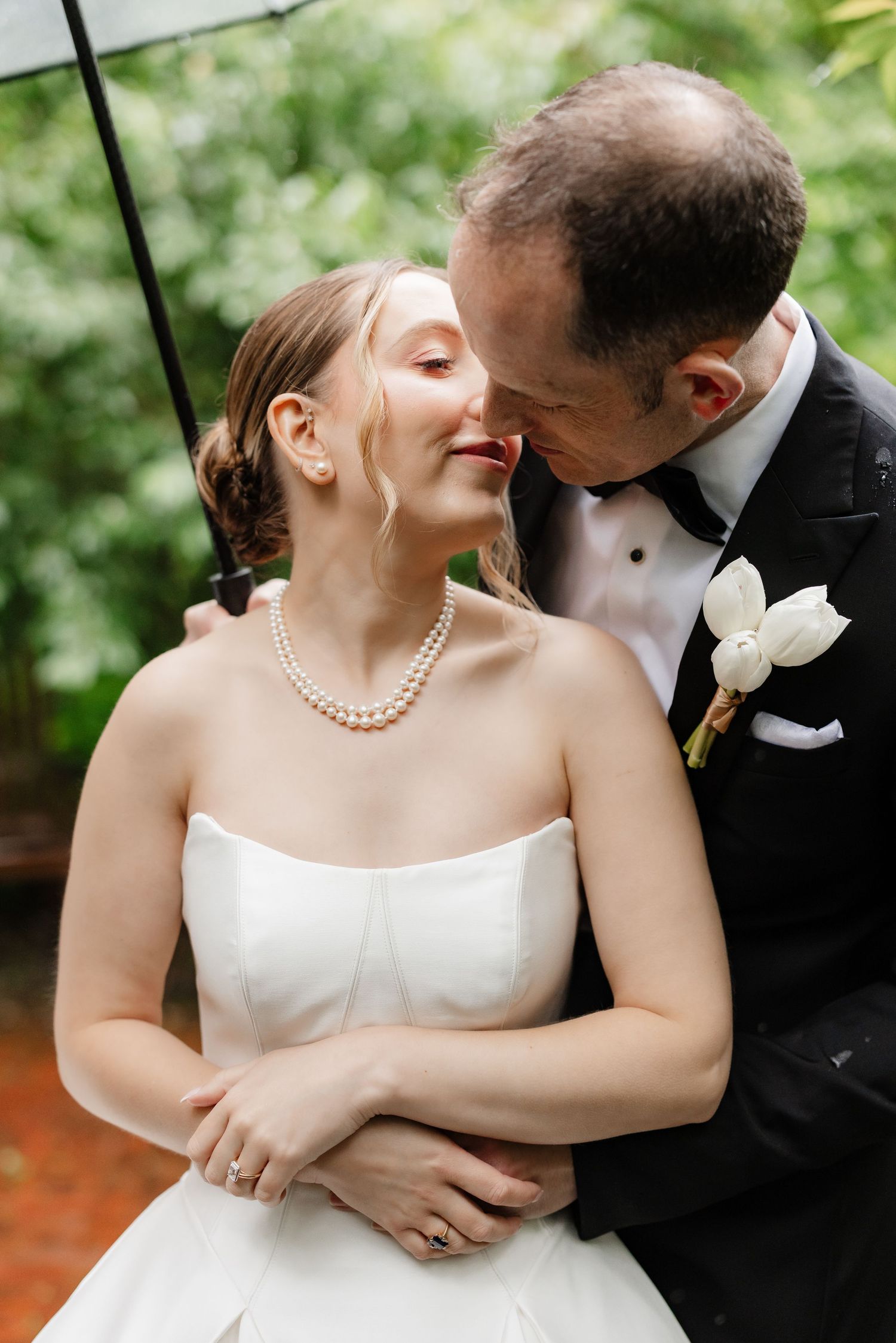 Groom in black tuxedo tenderly kisses bride wearing pearl necklace under a clear umbrella in lush greenery.