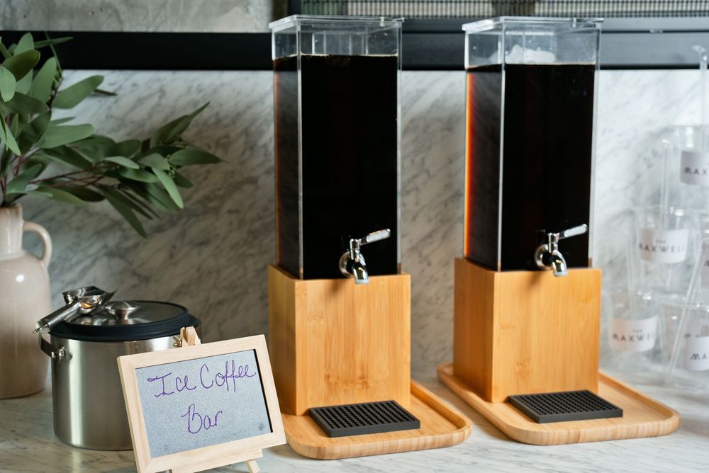 Two black beverage dispensers on bamboo stands at an ice coffee bar station on a marble countertop.
