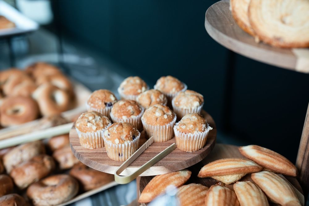 Assorted bakery display with muffins on a wooden stand, bagels, and madeleines at a cafe.