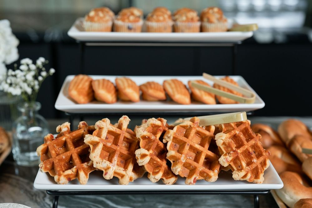 Freshly baked waffles and pastries displayed on white rectangular platters at a breakfast buffet spread.