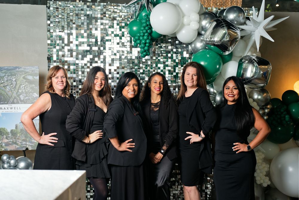Six women in black attire pose together at an elegant event with green, white, and silver balloon decor.