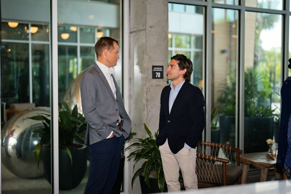 Two men in business casual attire having a conversation in a modern lobby with plants and glass doors.
