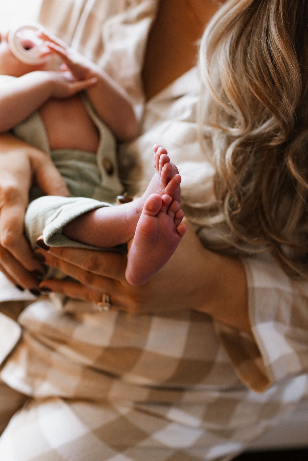 A mother in plaid pajamas gently holds her newborn baby, with tiny feet prominently displayed in a warm, intimate close-up.