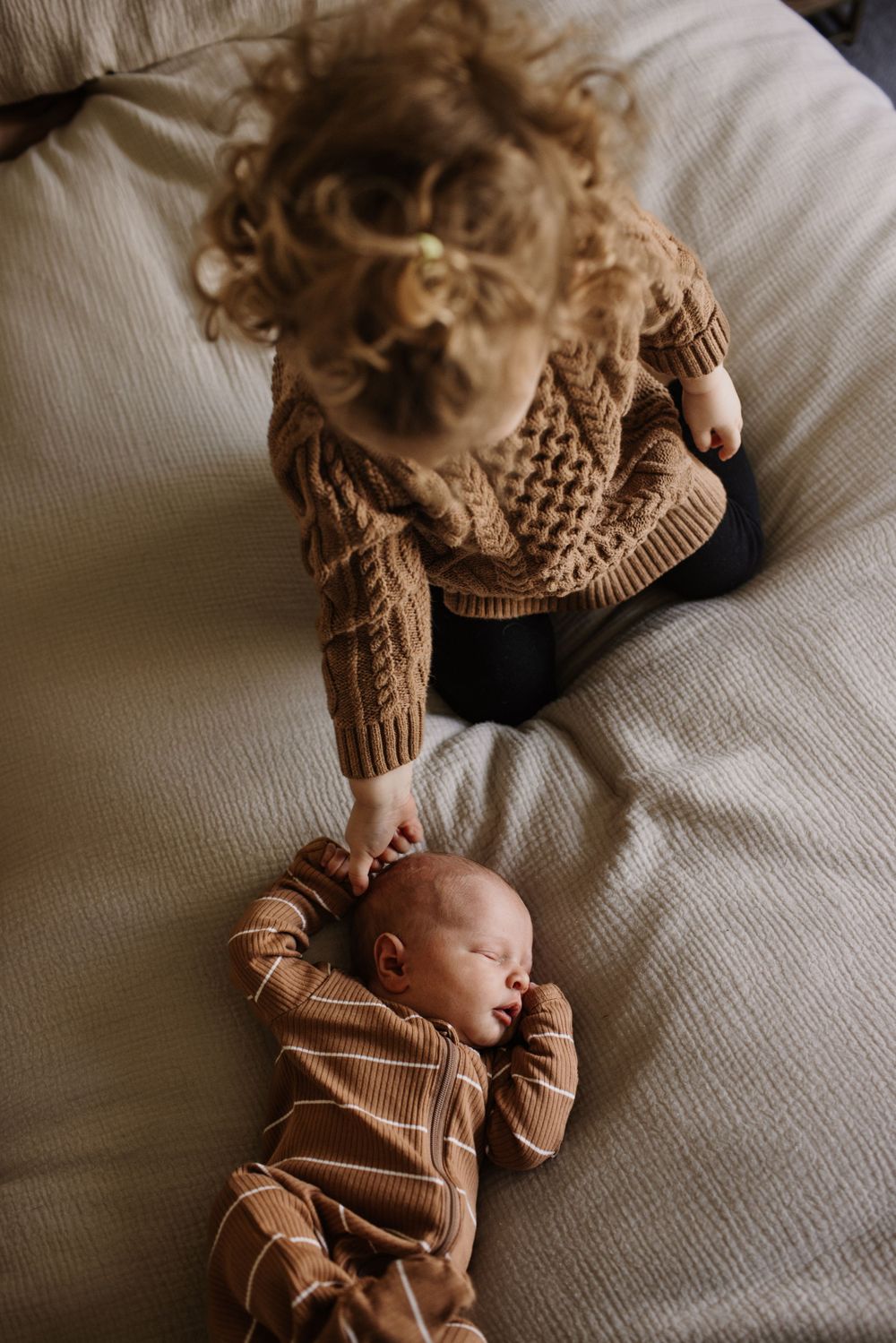 Young curly-haired toddler in a brown knit sweater gently touching a sleeping newborn sibling on a bed.