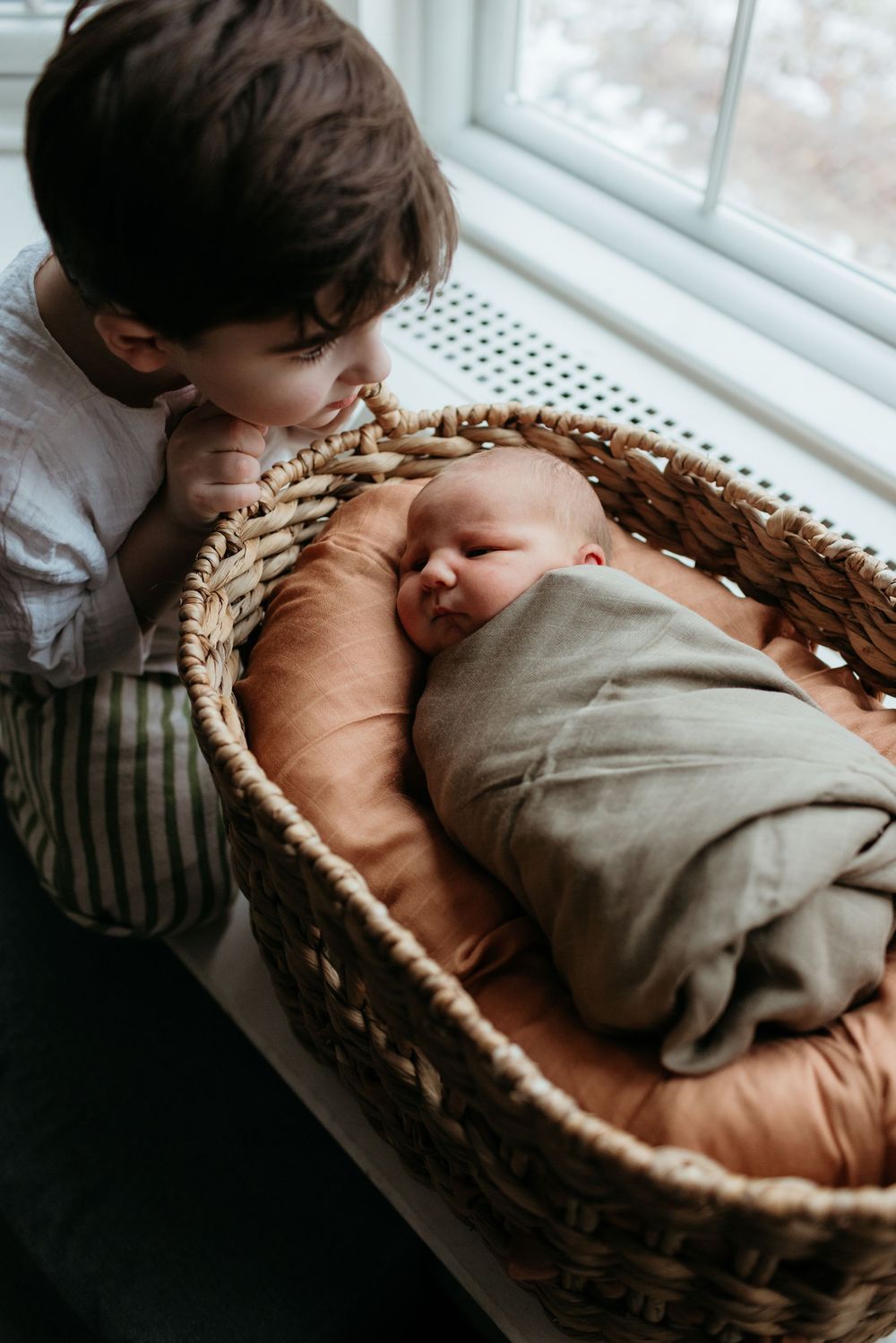 A young boy tenderly kisses a sleeping newborn baby wrapped in a gray swaddle inside a wicker Moses basket.
