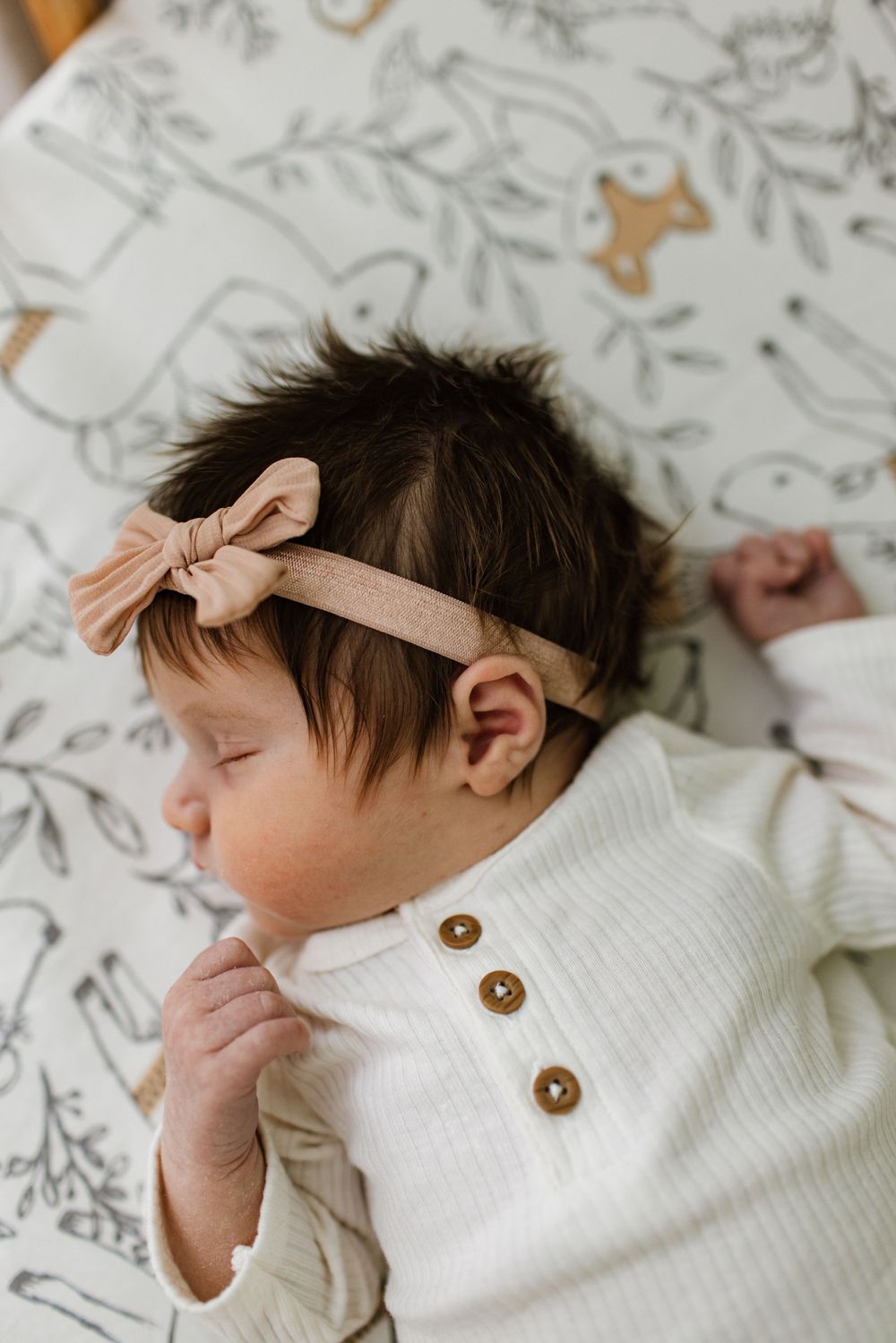 Close-up of a sleeping newborn baby girl with dark hair wearing a pink bow headband and white button onesie.