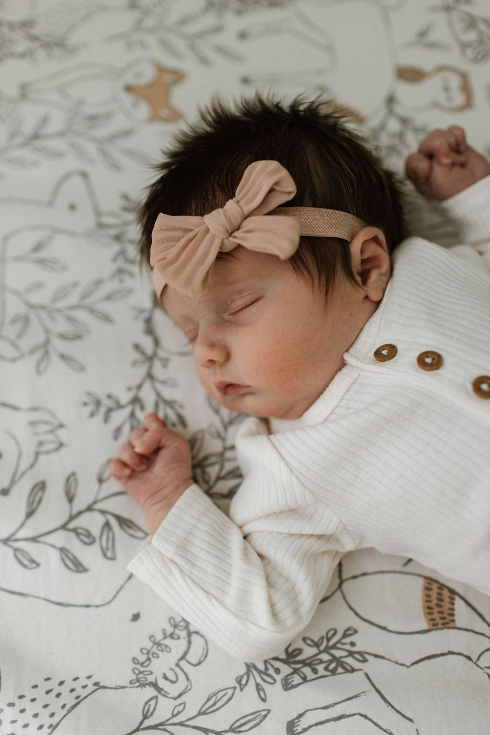 Sleeping newborn baby girl wearing a white ribbed onesie and pink bow headband on a floral patterned blanket.