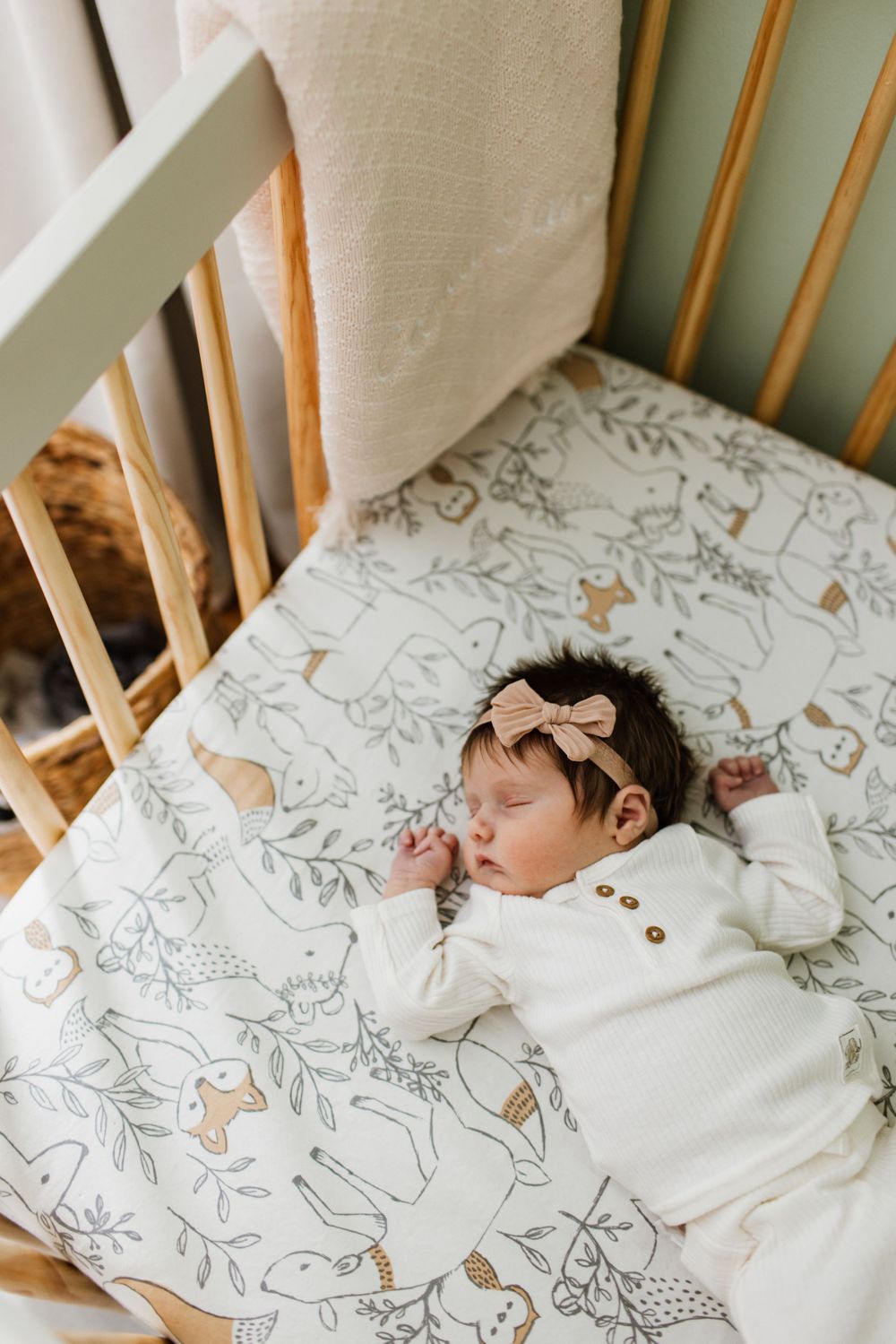 Newborn baby girl sleeping in a wooden crib on a woodland animal printed white sheet wearing a bow headband.