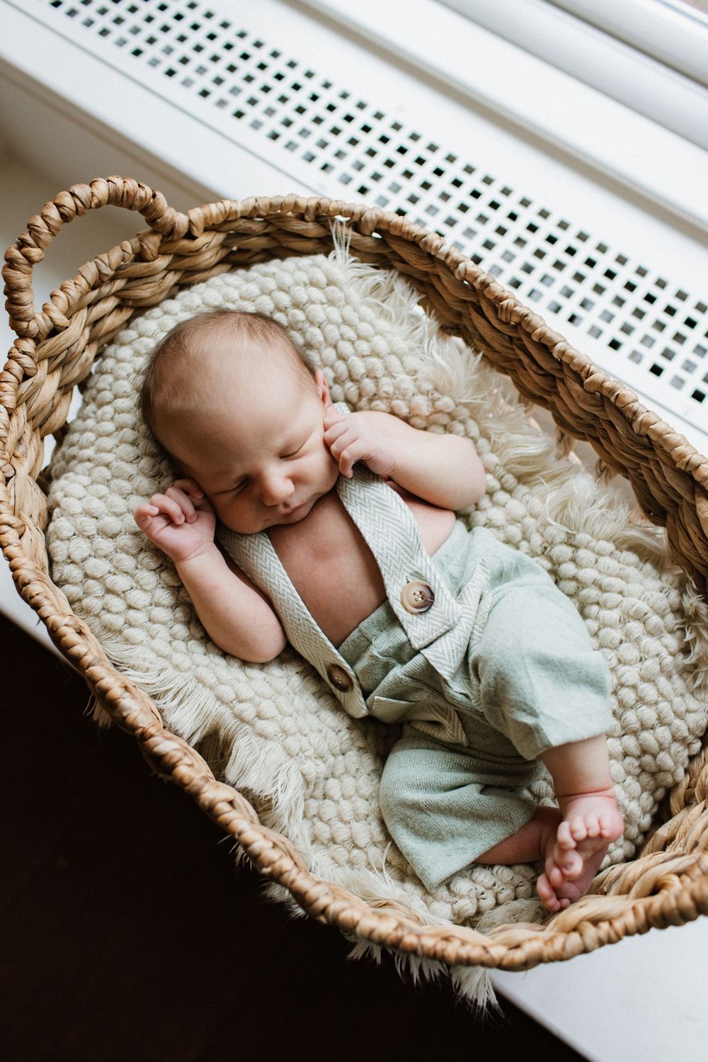Newborn baby sleeping peacefully in a woven Moses basket lined with cozy blankets, wearing sage green linen overalls.