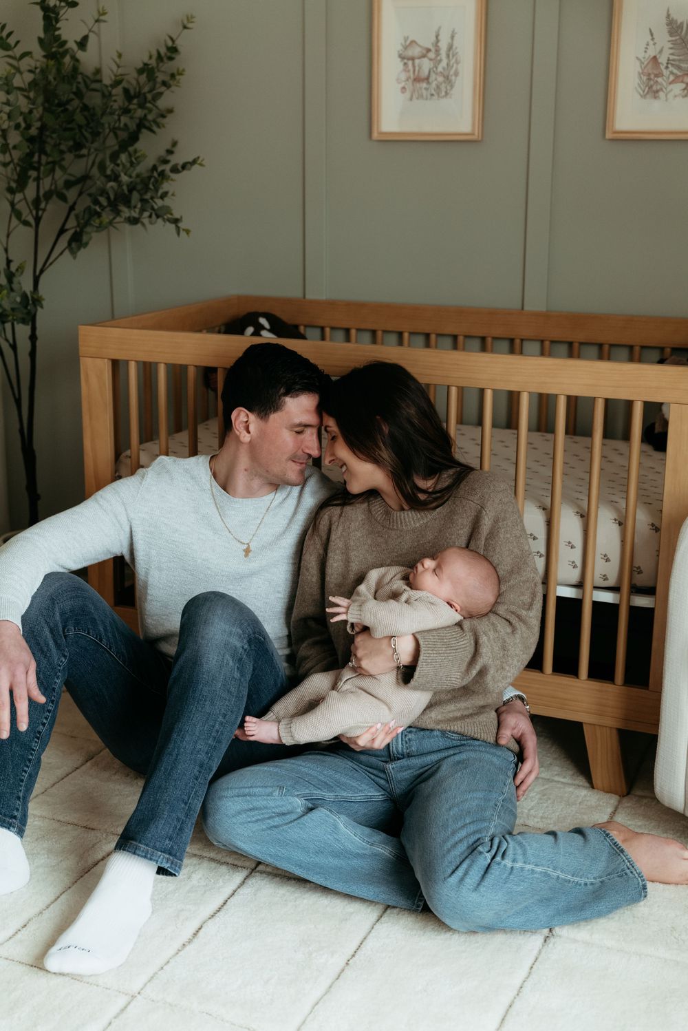 A couple sits on a nursery floor holding their newborn baby, leaning in for a kiss in front of a wooden crib.