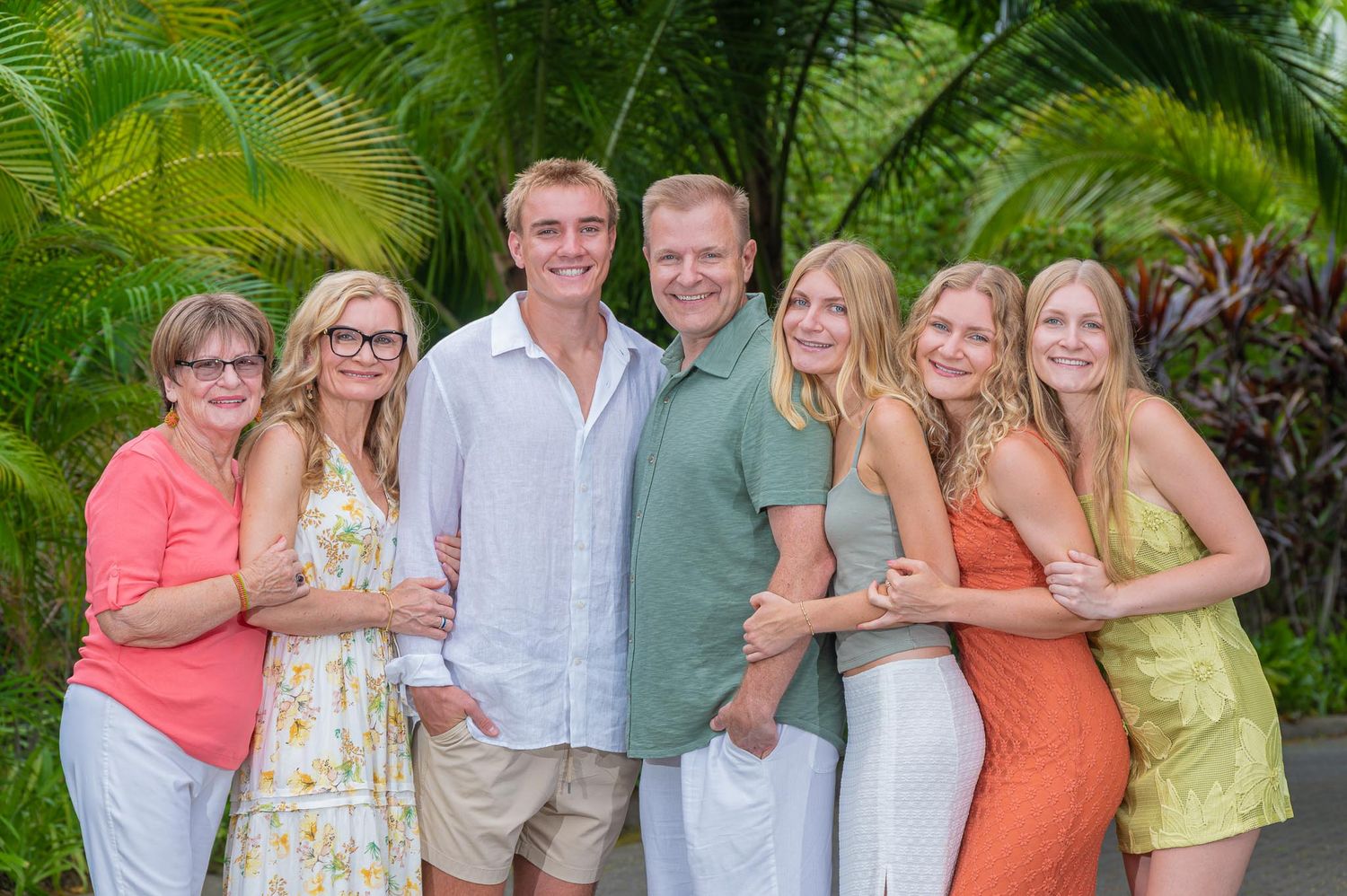 A cheerful family of seven poses together outdoors surrounded by lush tropical greenery in a warm vacation setting.