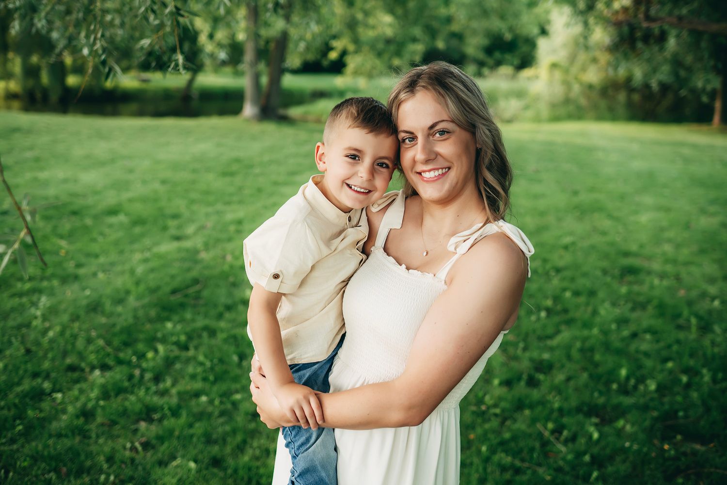 A smiling woman in a white dress holds a young boy in a beige shirt outdoors in a lush green park setting.