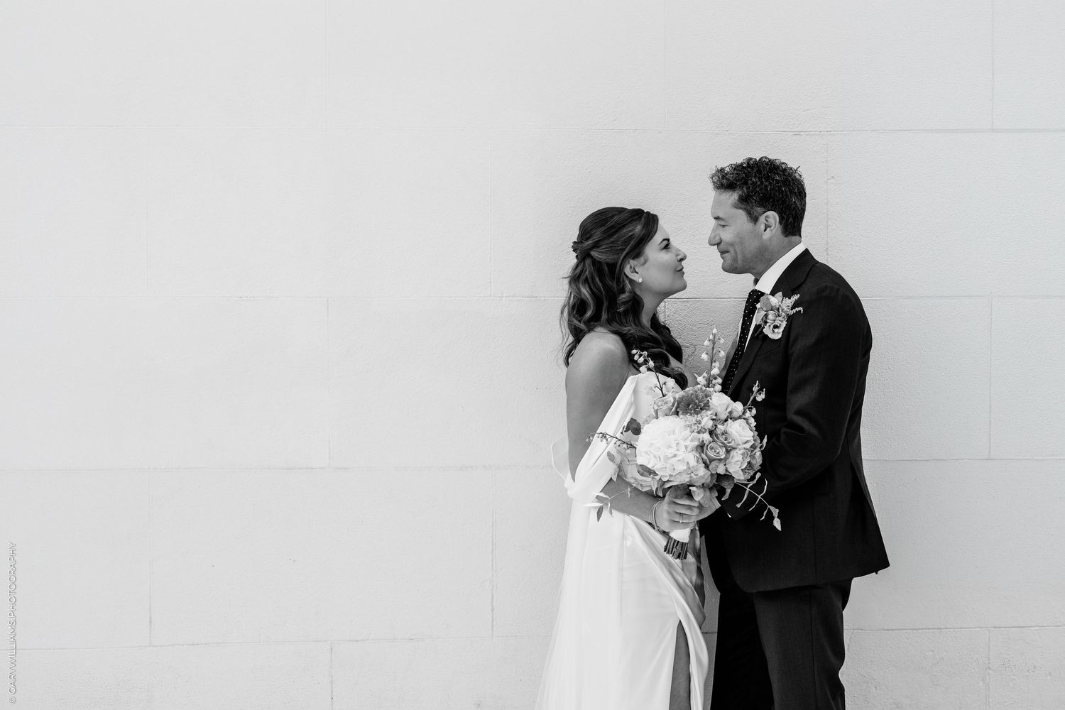 Black and white photo of a bride and groom sharing a romantic moment, bride holding a bouquet, against a white wall.