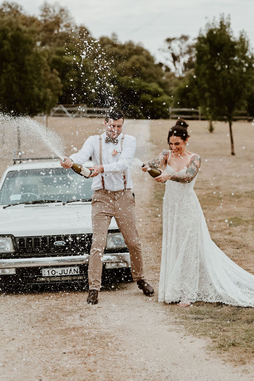 Bride and groom celebrating wedding by popping champagne bottles beside a vintage white truck outdoors.