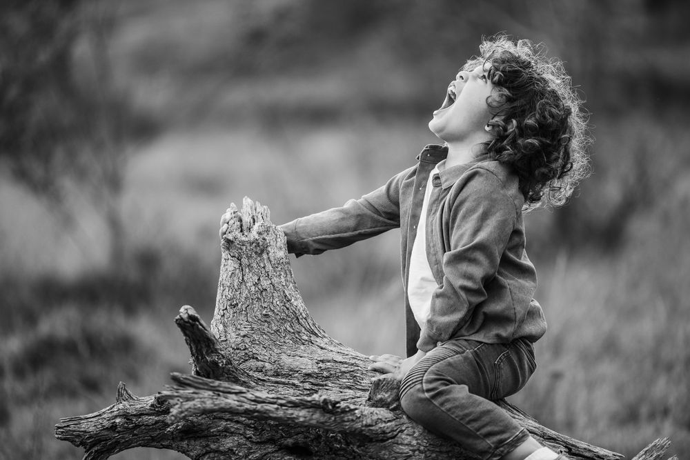 Black and white photo of a joyful curly-haired child sitting on a log, looking up with arms outstretched outdoors.