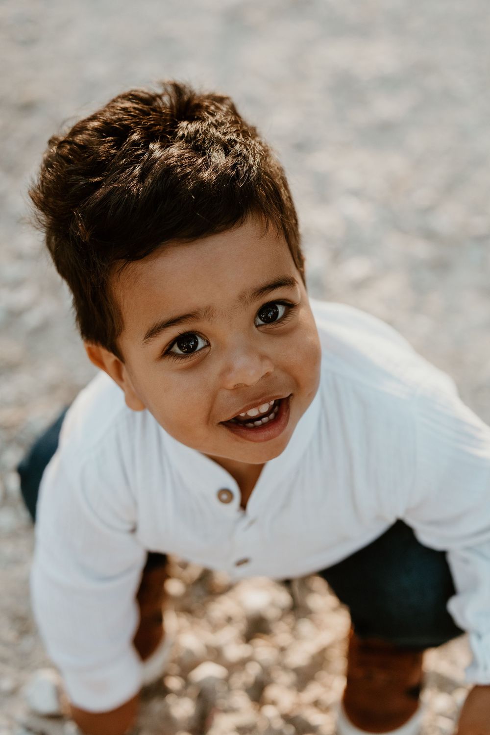A young boy in a white shirt smiles up at the camera while playing outdoors on a sunny day.