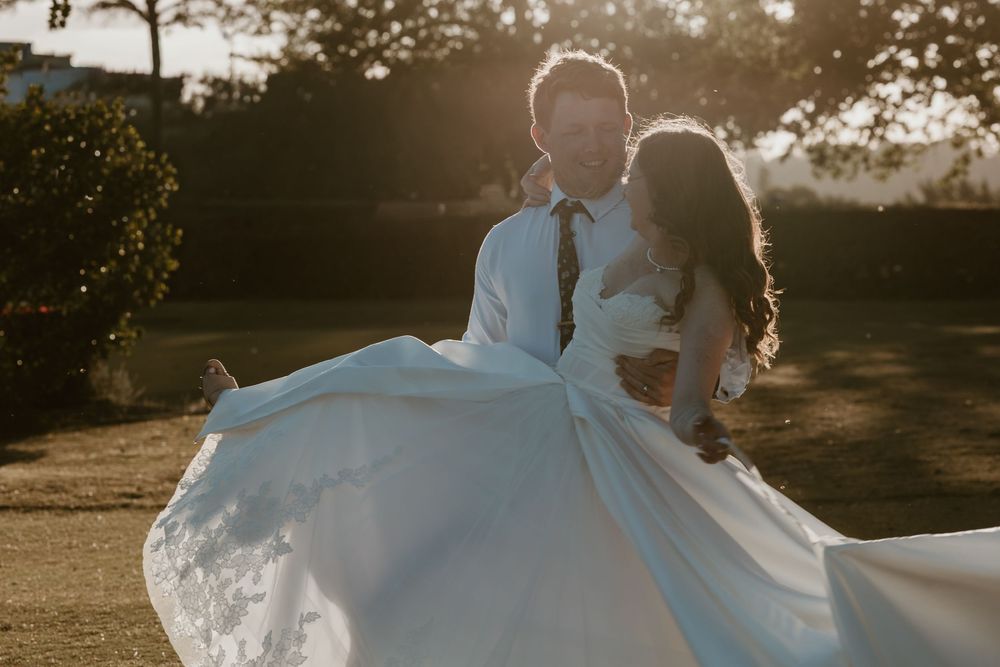 Couple sharing a romantic sunset kiss, bride's white gown flowing gracefully in golden hour light outdoors.