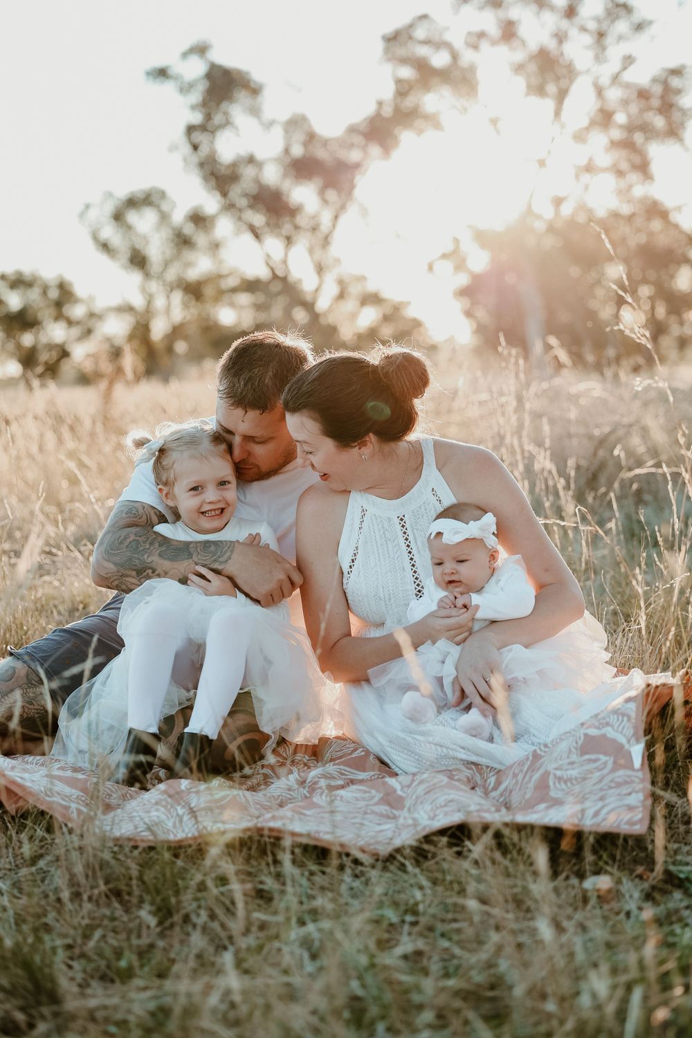 Family of four sitting on a blanket in a golden field at sunset, parents kissing their two young daughters.