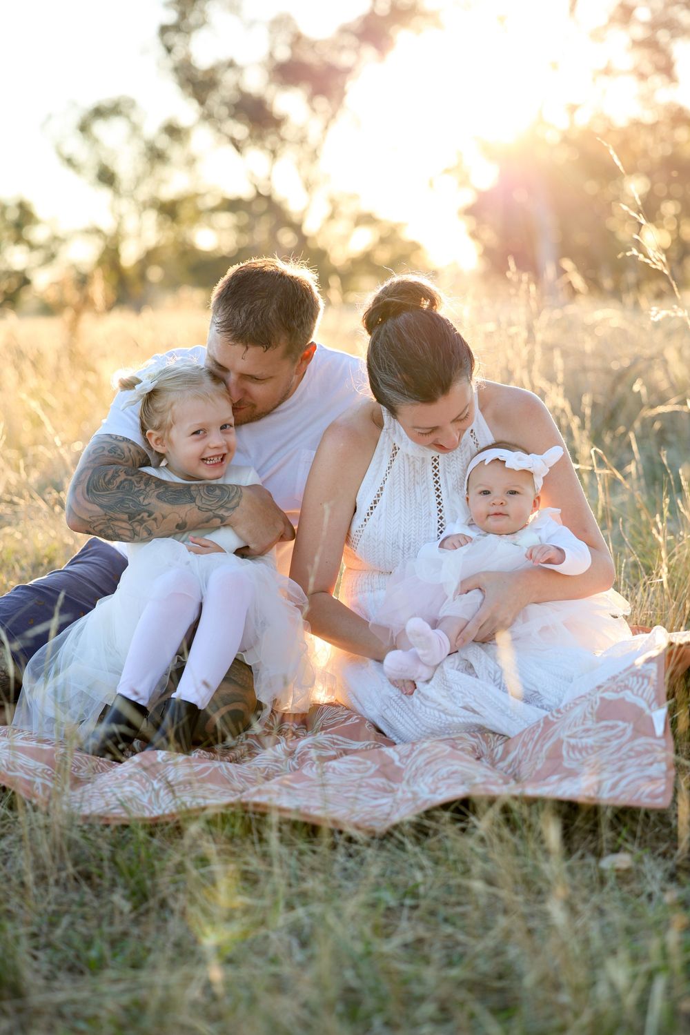 Family of four sitting on a blanket in a golden sunlit field, parents lovingly holding two young daughters dressed in white.