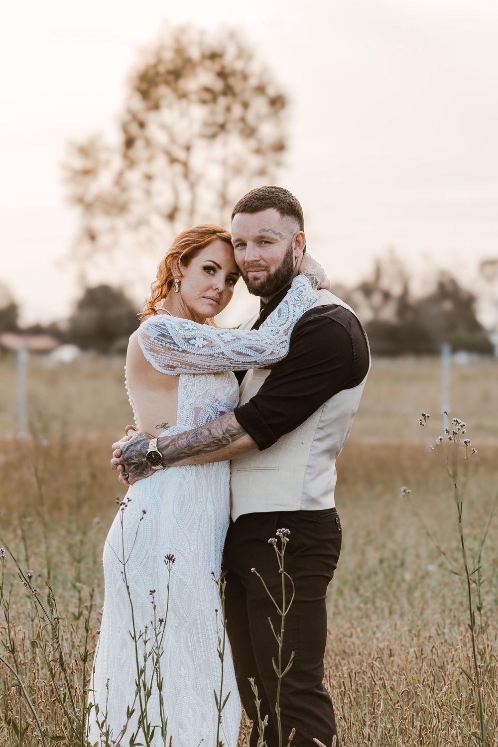 Couple embracing in a golden field at sunset, bride in white lace dress, groom in vest with tattoos.