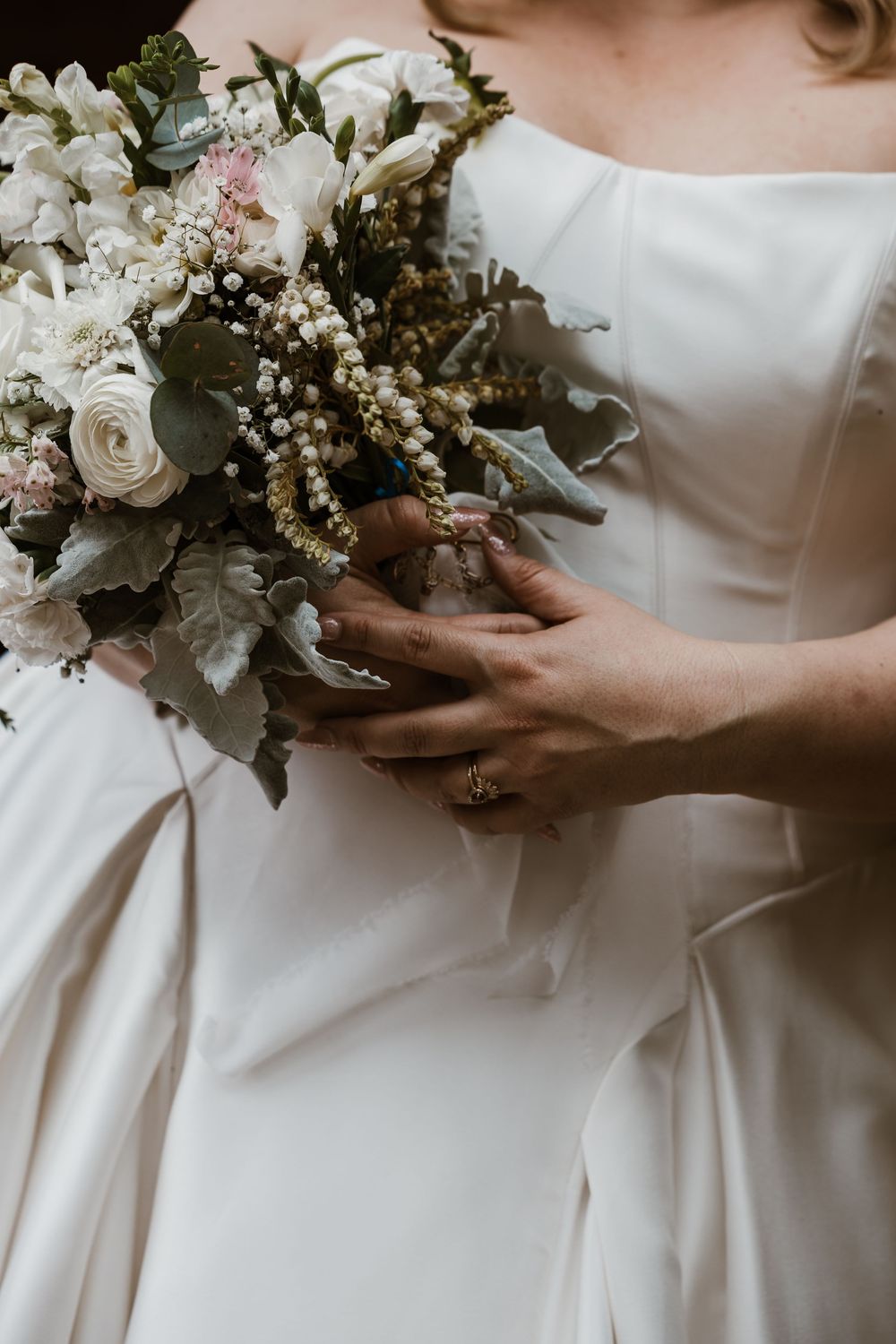 Bride in white gown holding elegant bouquet of white roses, greenery, and blush flowers, with gold ring visible on her hand.