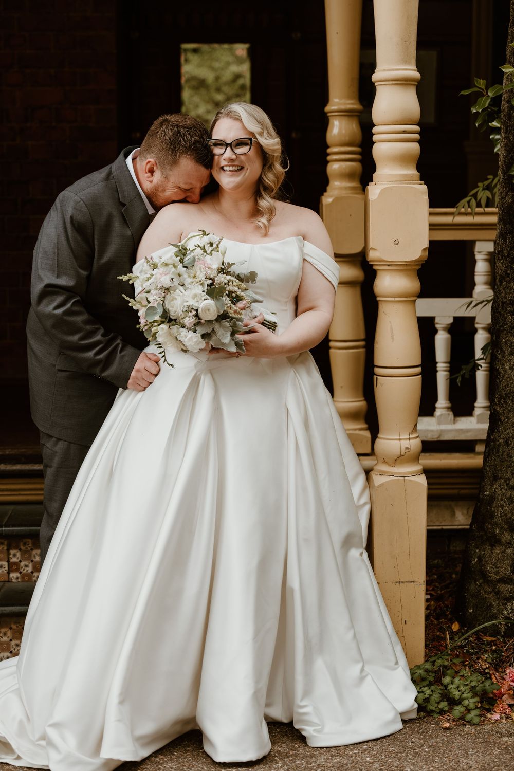 Bride in off-shoulder white ball gown holding bouquet while groom nuzzles her neck on a porch.