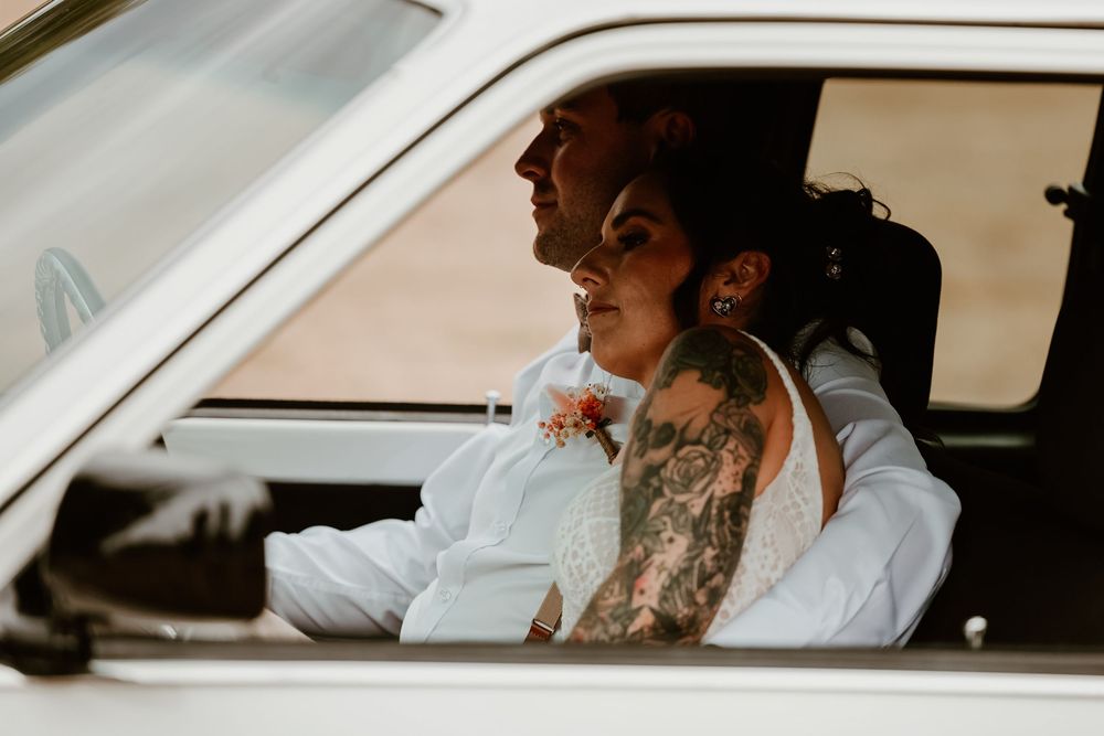A tattooed bride and groom share a tender moment inside a vintage white car on their wedding day.