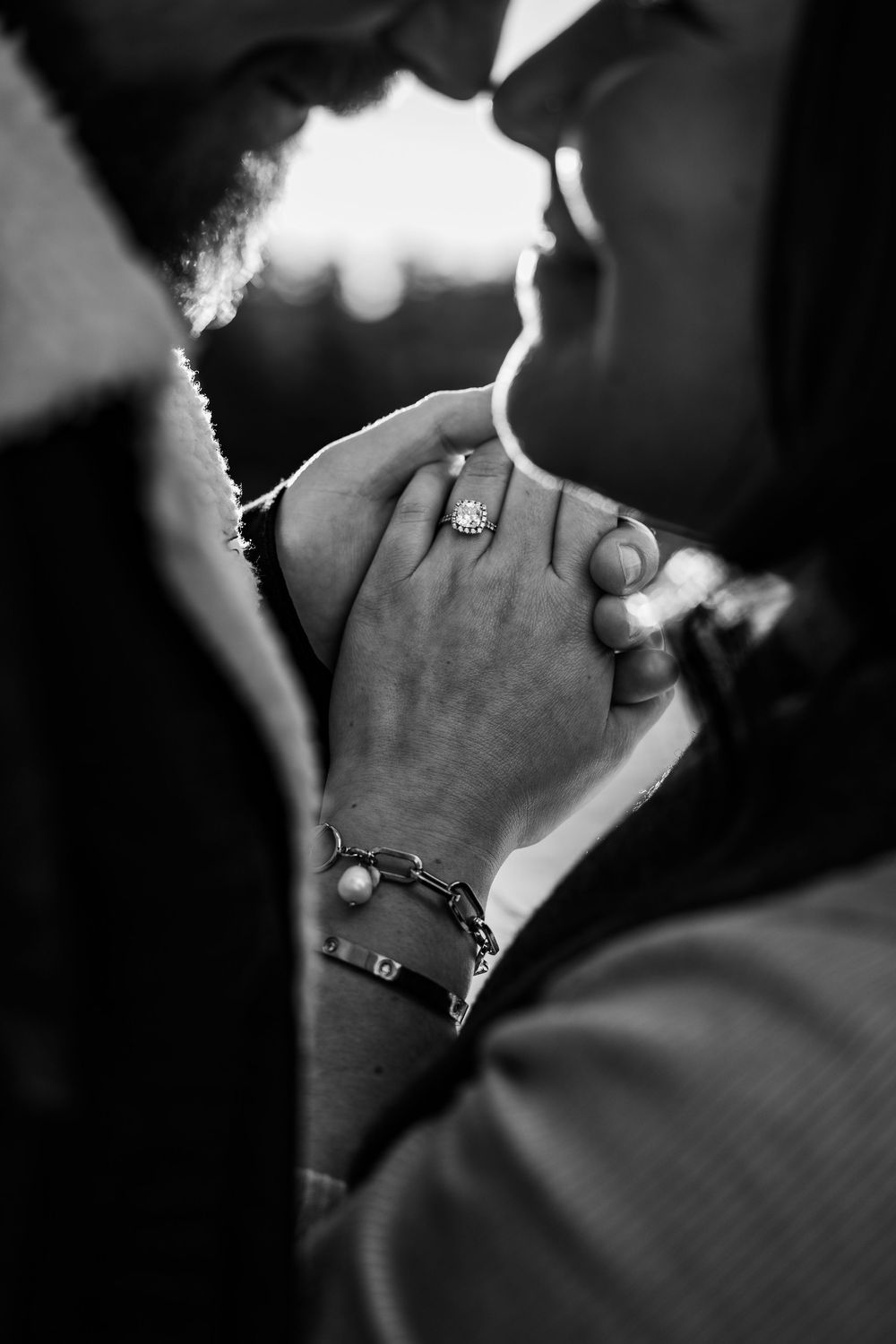 Black and white photo of couple nearly kissing, hands clasped showing diamond engagement ring and pearl bracelet.