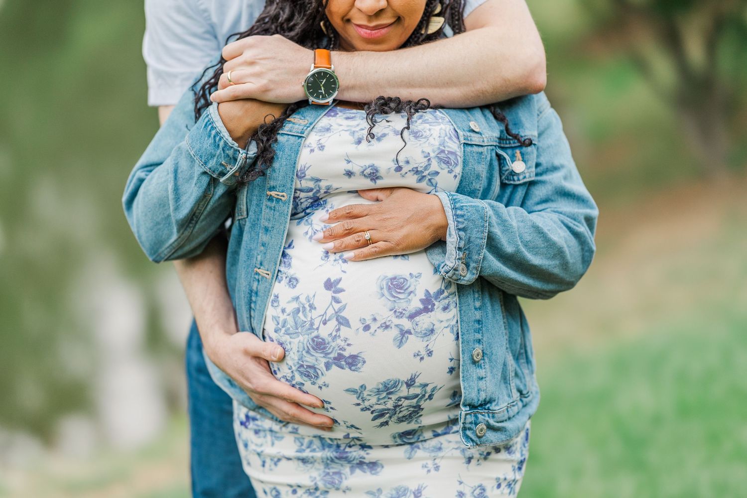 Pregnant woman in floral dress and denim jacket embraced by partner outdoors in a maternity photo session.