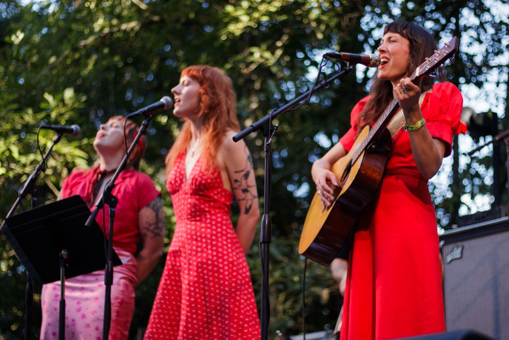 Three women in red dresses performing on stage, one playing acoustic guitar, singing into microphones outdoors.
